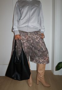 Henkilö, jolla on päällään harmaa Adidas-takki, camohousut, vaaleanbeiget korokepohjaiset saappaat, pitää käsissään suurta mustaa nahkakassia ja seisoo sisätilassa puulattialla.