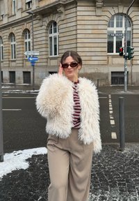 Femme portant une veste blanche duveteuse, un haut rayé et un pantalon beige ajuste ses lunettes de soleil dans une rue de la ville avec un bâtiment historique en pierre et des plaques de neige.