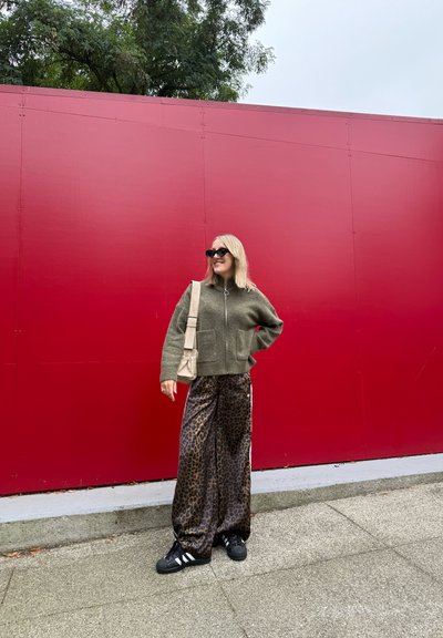 Chaqueta verde con cremallera, pantalones anchos de estampado de leopardo negro, zapatillas negras con rayas blancas y un bolso de hombro beige frente a una pared roja.