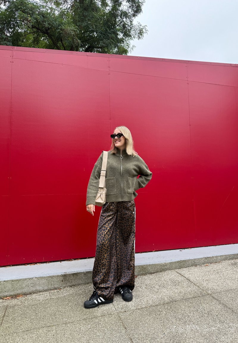 Veste zippée verte, pantalon large à motifs léopard noirs, baskets noires avec rayures blanches et un sac à épaule beige devant un mur rouge.