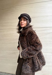 Manteau en fausse fourrure imprimé léopard avec grand col et fermetures par boutons, associé à un pull en maille crème et une jupe marron. Accessoire casquette noire.