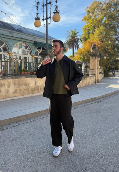 Hombre con chaqueta negra, camisa color oliva, pantalones negros y zapatillas blancas caminando por una calle pavimentada con árboles y un edificio al fondo.