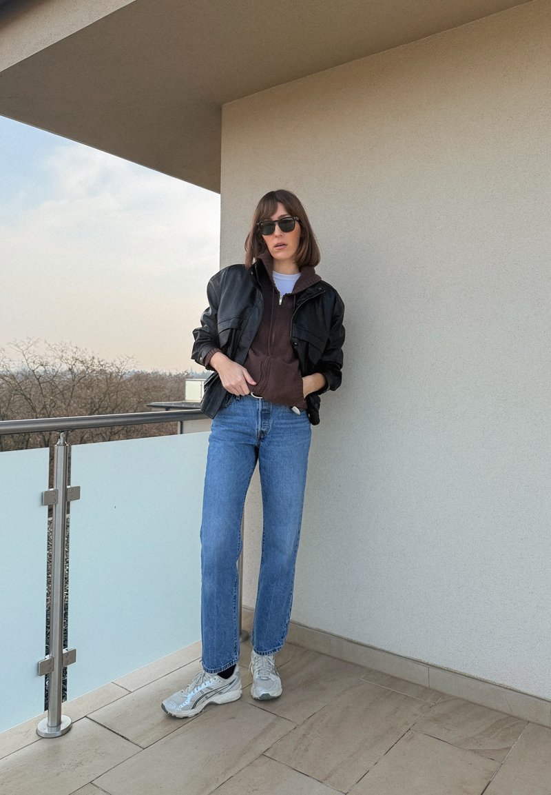Vrouw met zonnebril, zwarte leren jas, bruine hoodie, blauwe spijkerbroek en witte sneakers die op een balkon met mat glazen balustrade staat.