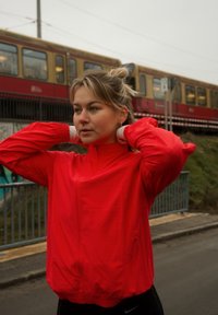 Jeune femme en veste rouge ajustant ses cheveux près des voies ferr ées avec un train en passage à l'arrière-plan par une journée nuageuse.