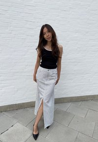 Νεαρή γυναίκα με μακριά μαλλιά στέκεται μπροστά σε λευκό τοίχο από τούβλα, φορώντας μαύρο αμάνικο τοπ, ριγέ μακριά φούστα με μπροστινό σκίσιμο και μαύρες γόβες.