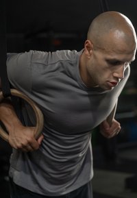 Αθλητής που εκτελεί muscle-up σε ξύλινες γυμναστικές ρινγκ, φορώντας κοντομάνικο γκρι μπλουζάκι, δείχνοντας μυϊκή ανάπτυξη και συγκέντρωση.