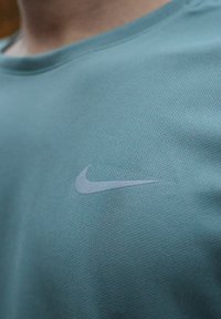 Un t-shirt Nike turquoise avec un logo Nike gris clair. Le tissu présente un design en maille texturée et respirante.