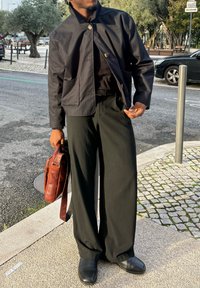 Uomo con camicia nera, giacca nera, pantaloni larghi neri e scarpe nere, che tiene una borsa in pelle marrone, in piedi sul marciapiede urbano vicino ad auto parcheggiate.