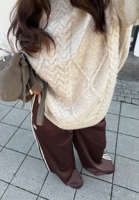Kraftig cremefarvet kabelstrikket sweater, brune satin atletikbukser med hvide striber og en grå ruskindstaske, placeret mod en flisebelagt overflade.