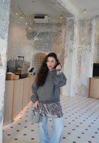 Femme aux longs cheveux foncés portant des lunettes, un sweat à capuche gris pailleté, une jupe à sequins par-dessus un jean, et tenant un petit sac à main gris dans un café rustique.