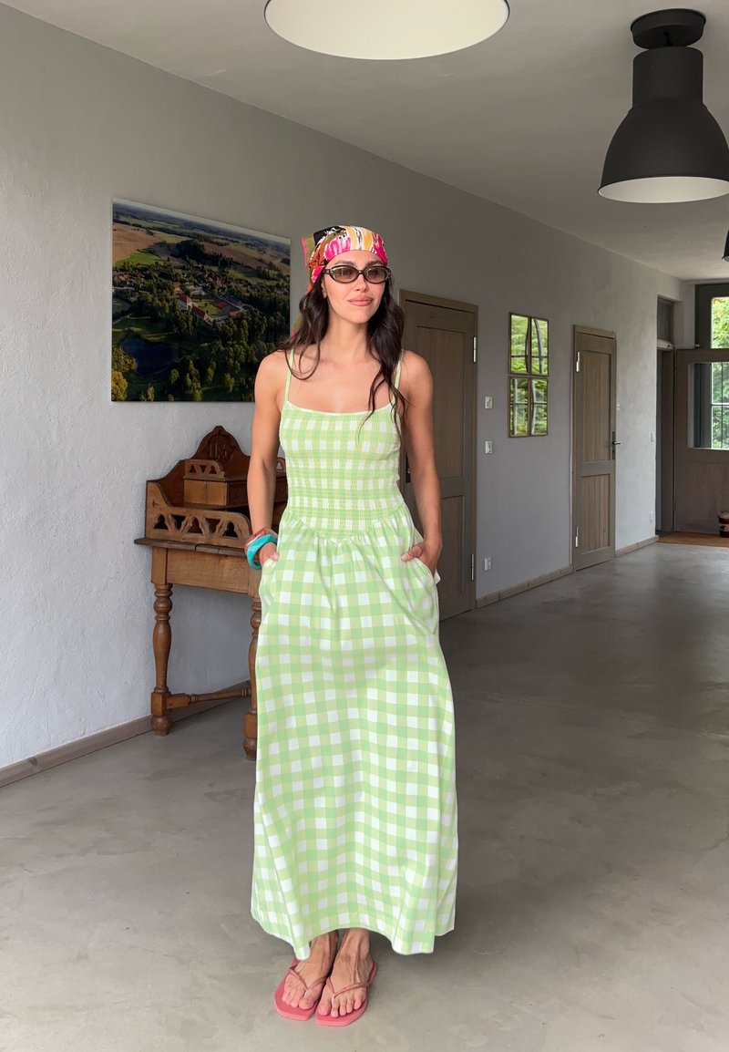 Femme portant une robe à carreaux verts et blancs, un bandeau coloré, des lunettes de soleil et des tongs roses, se tenant dans un couloir lumineux avec des portes en bois.