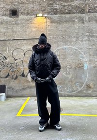 Fekete bélelt dzseki szőrmével bélelt kapucnival, fekete nadrággal párosítva. Kiegészítők közé tartoznak a fekete kesztyűk és sapka, melyek egy texturált fal előtt láthatók.