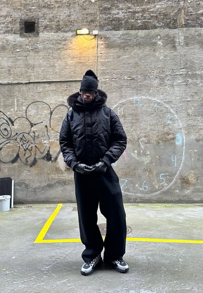 Chaqueta negra acolchada con capucha forrada de piel, combinada con pantalones negros. Complementos incluyen guantes negros y un gorro, sobre un fondo de pared texturizada.