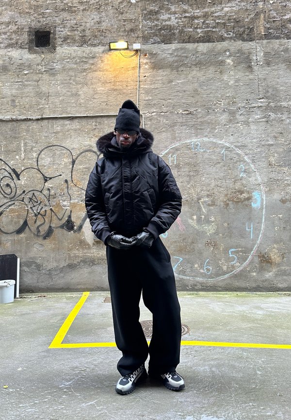 Chaqueta negra acolchada con capucha forrada de piel, combinada con pantalones negros. Complementos incluyen guantes negros y un gorro, sobre un fondo de pared texturizada.