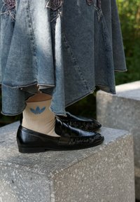 Μαύρες δερμάτινες loafers με υφή, συνδυασμένες με κρεμ κάλτσες με ένα ανοιχτό μπλε λογότυπο Adidas, φορεμένες με τζιν φούστα.