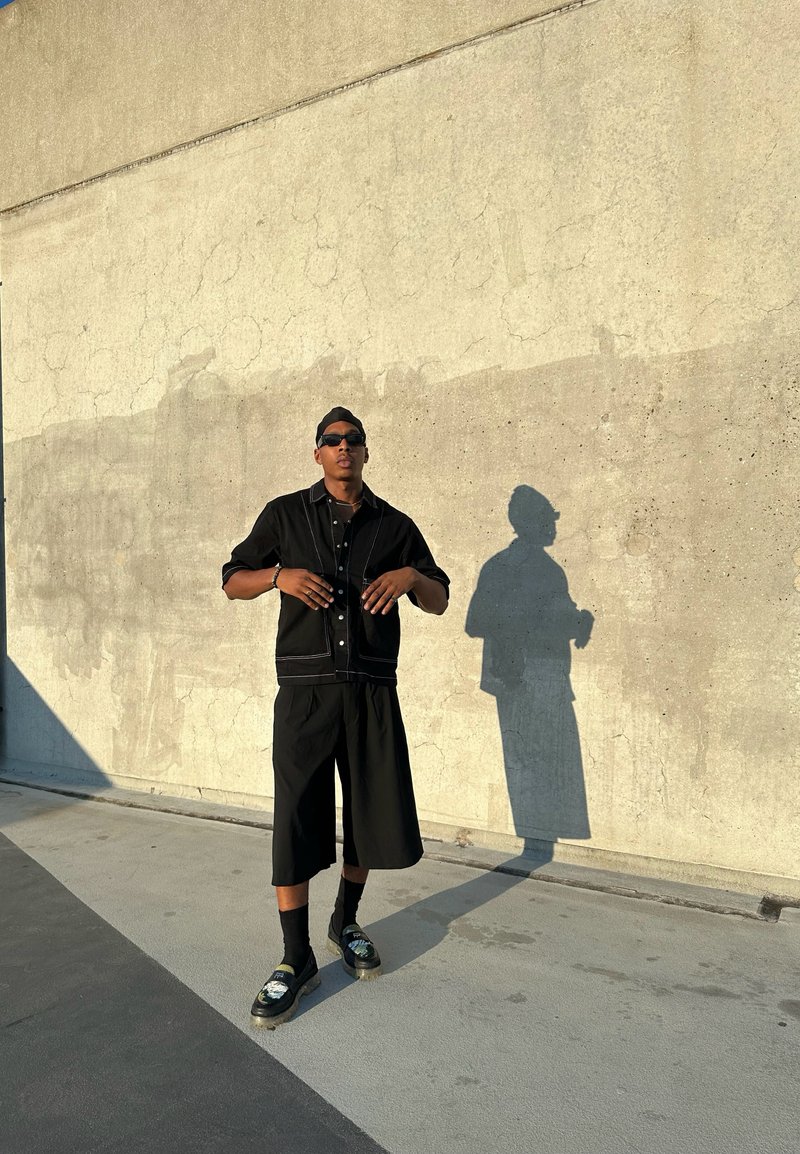 Μαύρο oversized πουκάμισο με λευκή ραφή, αντίστοιχες χαλαρές κοντές παντελόνες και σκούρα παπούτσια με γραφικό σχέδιο, σε φόντο από υφή σκυρόδεμα.