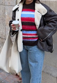 Personne portant une veste en cuir doublée de shearling, un pull rayé rouge et bleu, un jean bleu, tenant une tasse de café et un grand sac cabas crème.
