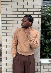 Sweat-shirt zippé marron clair avec un col haut, associé à un pantalon plissé marron. Le modèle porte des lunettes transparentes et des bijoux. Fond mur de briques.