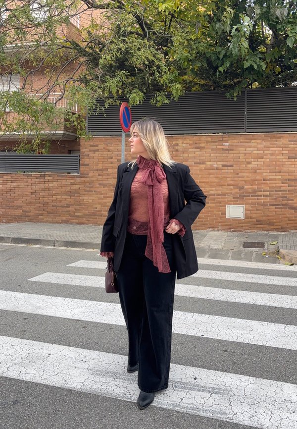 Femeie îmbrăcată într-un sacou și pantaloni negri, traversând strada pe trecerea pentru pietoni, uitându-se în lateral, ținând o geantă mică mov.