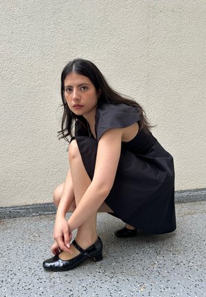 Joven mujer con cabello largo y oscuro, vestida con un vestido negro y zapatos negros, agachada junto a una pared clara con textura y suelo moteado.