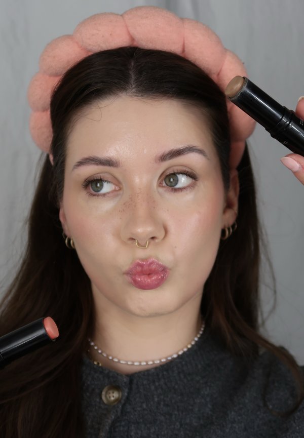 Kvinne med rosa hårbånd som holder to sminkestifter nær ansiktet, smiler og ser til siden.