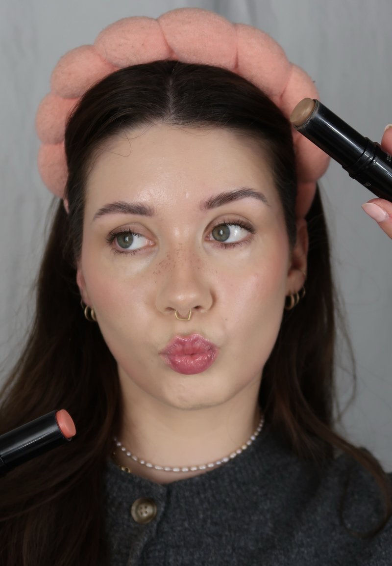 Femme avec un bandeau rose tenant deux bâtonnets de maquillage près du visage, les lèvres en cul-de-poule et regardant sur le côté.