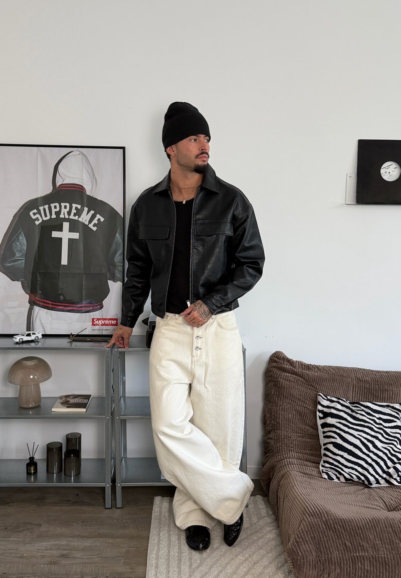 Homme portant un bonnet noir, une veste en cuir noire et un pantalon blanc large, debout à côté d'une étagère métallique avec des objets d’art et de décoration dans un salon minimaliste.