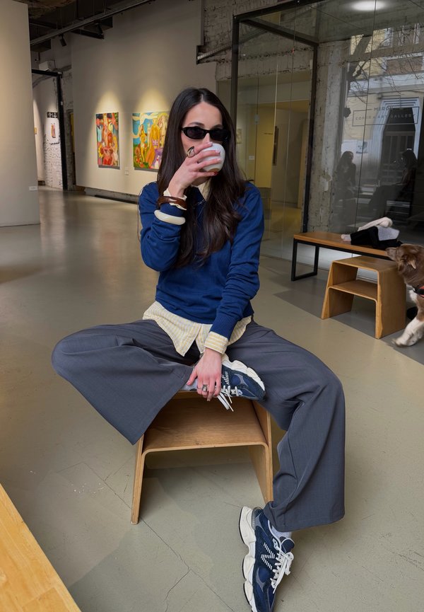 Žena v slnečných okuliaroch sedí na stoličke so založenými nohami v galérii umenia, popíja z pohára, vedľa nej je pes a na stene sú obrazy.