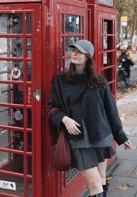 Mladi žena u sivoj kapici, tamnoj jakni i suknji naslonjena na crvenu britansku telefonsku govornicu na pločniku prekrivenom lišćem u gradu.