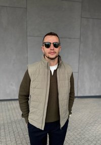 Bărbat purtând ochelari de soare, vestă bej matlasată și pulover olive, stând cu mâinile în buzunare în fața unui perete gri placat cu faianță.