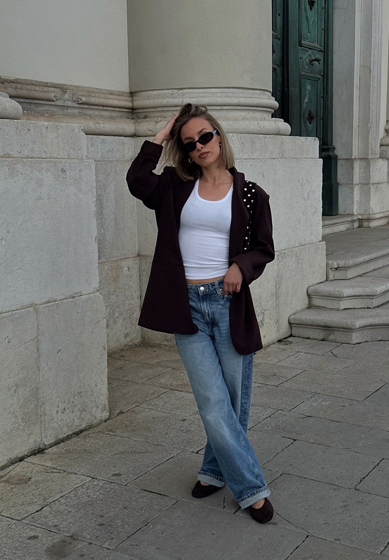 Femme portant un blazer noir, un débardeur blanc, un jean bleu et des lunettes de soleil, posant devant un immeuble en pierre avec des marches sur un trottoir pavé.