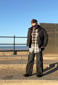 Veste matelassée noire par-dessus une chemise grise à carreaux et un pantalon cargo noir avec poches. Lunettes de soleil noires et chaussures à semelles épaisses. Plage en arrière-plan.