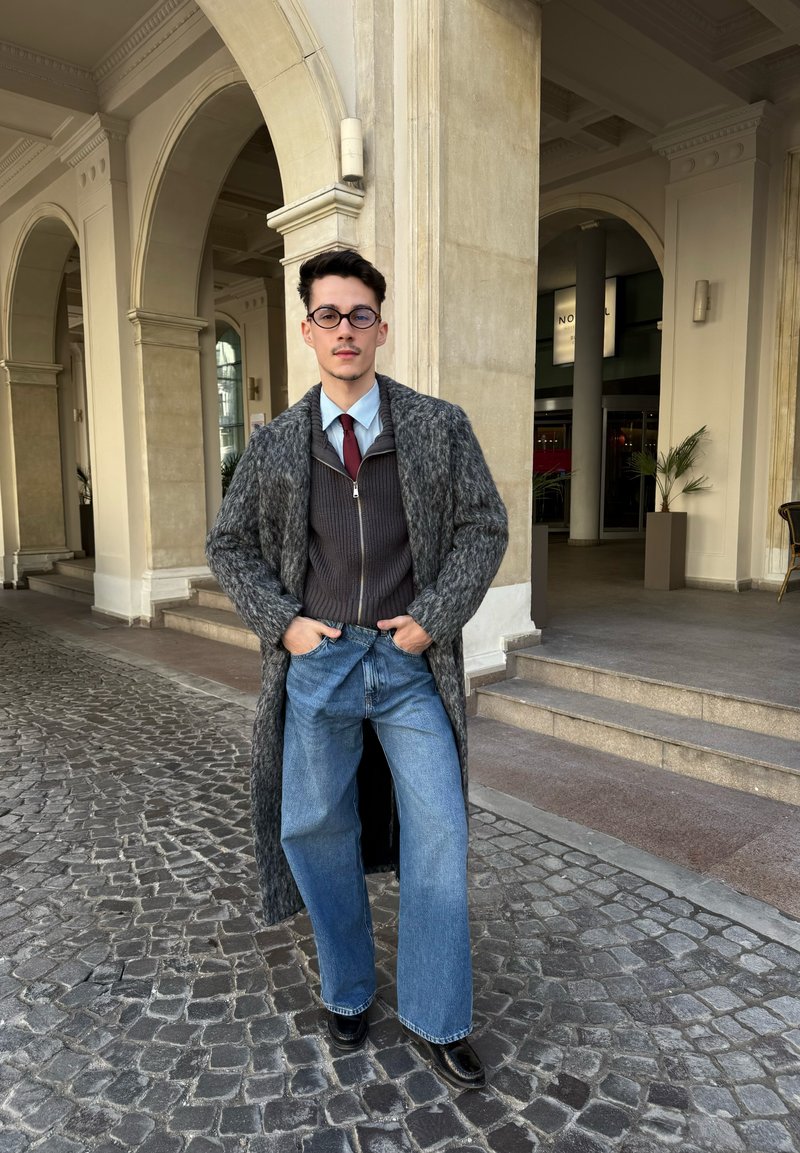 Νέος άνδρας με γυαλιά που φορά μακρύ γκρι παλτό, ζακέτα με φερμουάρ, κόκκινη γραβάτα, μπλε τζιν και μαύρα παπούτσια, στέκεται σε δρόμο με λιθόστρωτο.