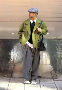 Groene wollen peacoat met grote knopen, zwarte gebreide trui, witte blouse met kraag, grijze brede broek en metallic sneakers. Zwarte tas en pet.