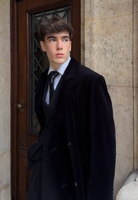 Une personne portant un manteau en velours foncé se tient à côté d'une porte en bois, vêtue d'un costume noir avec une cravate, et une chemise bleu clair. Texture douce, boutons visibles.