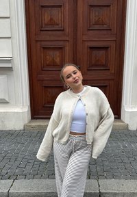 Jasno beżowy dziergany kardigan z szerokimi rękawami, biały krótki top i szare spodnie w paski. W tle znajduje się drzwi z panelami drewnianymi.