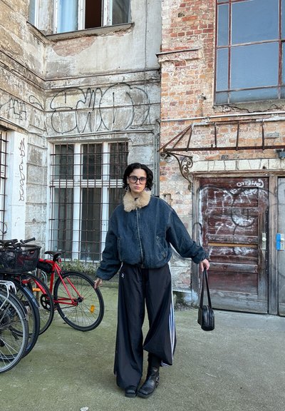 Persona con chaqueta vaquera de gran tamaño con cuello de piel y pantalones anchos negros de pie cerca de bicicletas y paredes de ladrillo cubiertas de grafitis.