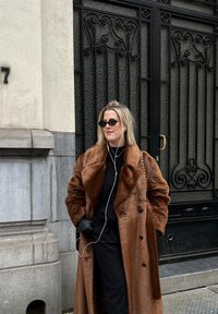 Γυναίκα που φοράει μακρύ καφέ γούνινο παλτό και μαύρα γυαλιά ηλίου στέκεται με τα χέρια στις τσέπες κοντά σε μια περίτεχνη μαύρη μεταλλική πύλη, ακούγοντας με ακουστικά.