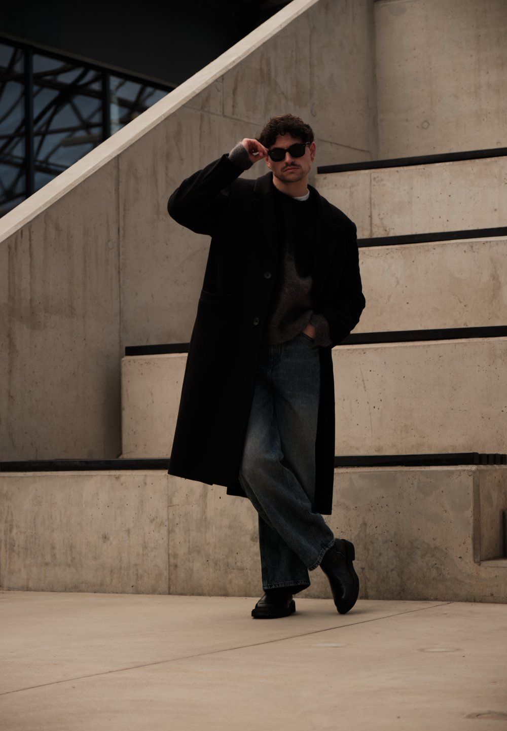 Homme portant un manteau noir, des lunettes de soleil et un jean, appuyé contre des marches en béton, une main dans la poche et l'autre ajustant ses lunettes.