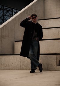 Homme portant un manteau noir, des lunettes de soleil et un jean, appuyé contre des marches en béton, une main dans la poche et l'autre ajustant ses lunettes.