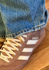 Brązowy zamszowy but sportowy z białymi skórzanymi paskami, wyposażony w białe sznurowadła i gumową podeszwę, zestawiony z jasnoniebieskimi, postrzępionymi dżinsami.