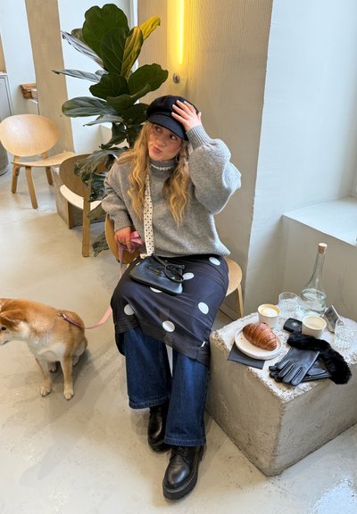 Mujer con suéter gris y gorra negra sentada en interiores con un perro Shiba Inu con correa, y una mesa cercana con café, un croissant y guantes.