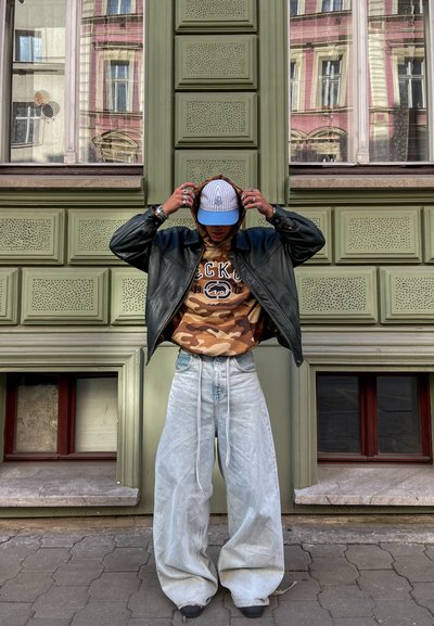 Chaqueta de cuero negra, sudadera de camuflaje, vaqueros de gran tamaño en azul claro y una gorra blanca con acentos marrones. Anillos plateados en las manos. Fondo de pared verde.