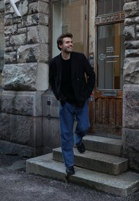 Pessoa vestindo um blazer preto sobre uma camisa preta e jeans azuis folgados, descendo degraus de pedra ao lado de uma porta de madeira e uma parede de pedra texturizada.