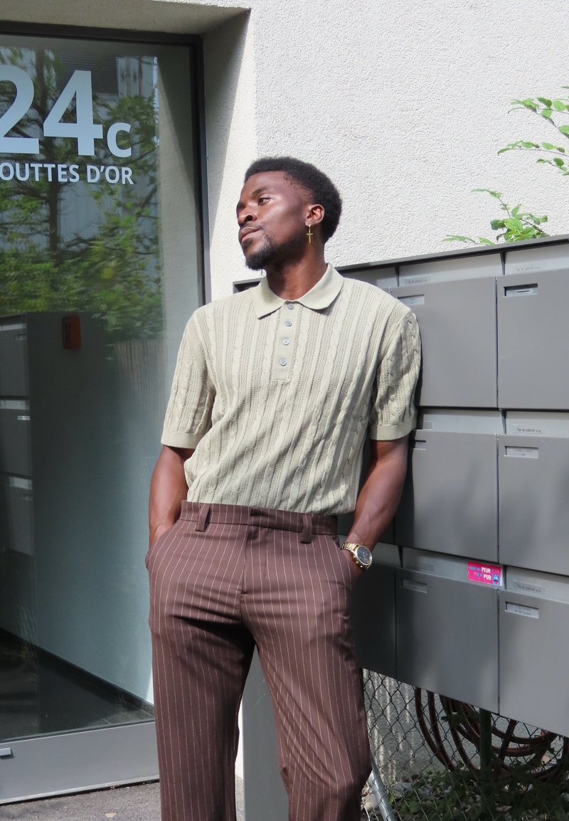 Polo shirt tricoté de couleur vert clair à manches courtes, associé à un pantalon marron à rayures. Montre en or et boucles d'oreilles en tant qu'accessoires notables.