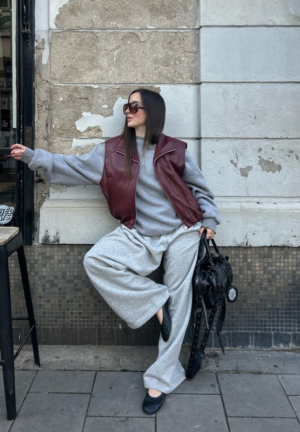 Woman in oversized grey sweatpants and sweater with burgundy vest, holding a black bag, posing by a worn stone wall on a city sidewalk.