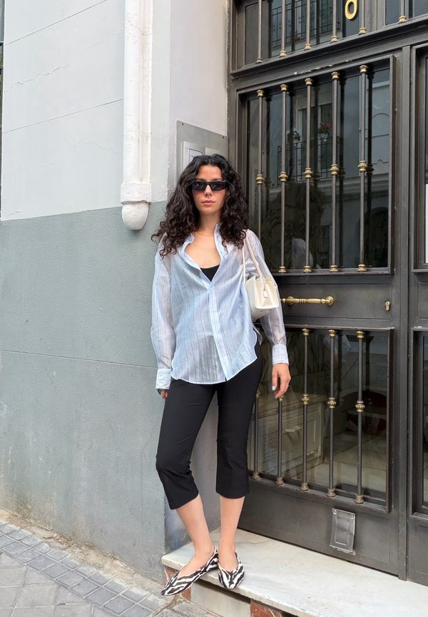 Donna con capelli ricci che indossa occhiali da sole, camicia bianca a righe, pantaloni neri corti e ballerine con stampa zebra, in piedi accanto a una porta in metallo nero in una strada cittadina.