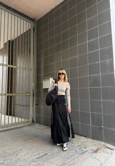 Falda maxi negra con volantes y franjas laterales blancas, combinada con una blusa ajustada beige sin hombros. Los accesorios incluyen un bolso negro y gafas de sol.