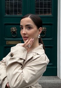 Beige trenchcoat med stor krave, knapdetaljer og optrukne ærmer. Sølv hoops øreringe og lagde ringe komplementerer outfittet. Grøn dør som baggrund.