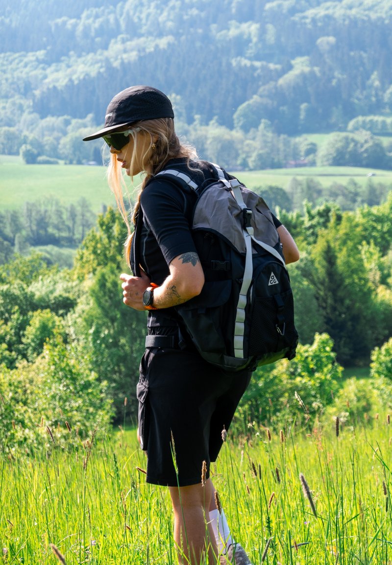 Femme portant une casquette et des lunettes de soleil fait de la randonnée avec un sac à dos à travers un champ herbeux, entourée d’arbres verts et de collines au loin.